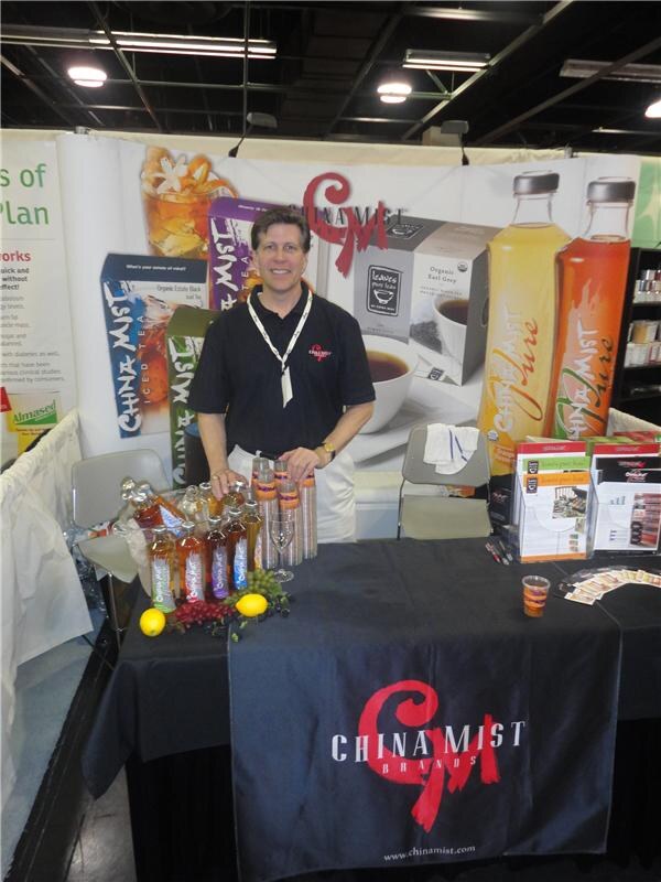 A smiling man in a black China Mist polo stands behind a black-draped table with bottles of iced tea, lemons and grapes. Behind him are China Mist boxes and bottles on a banner.