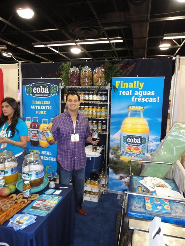 A smiling man in a purple plaid shirt holds two bottles in a Coba Aguas Frescas booth with banners reading 'Finally real aguas frescas!'. A woman in a blue shirt stands at left; jars sit on a table.