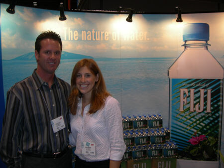 A man and a woman with name badges smile in front of a Fiji Water backdrop reading "The nature of water," with a large Fiji bottle graphic and stacked cases.