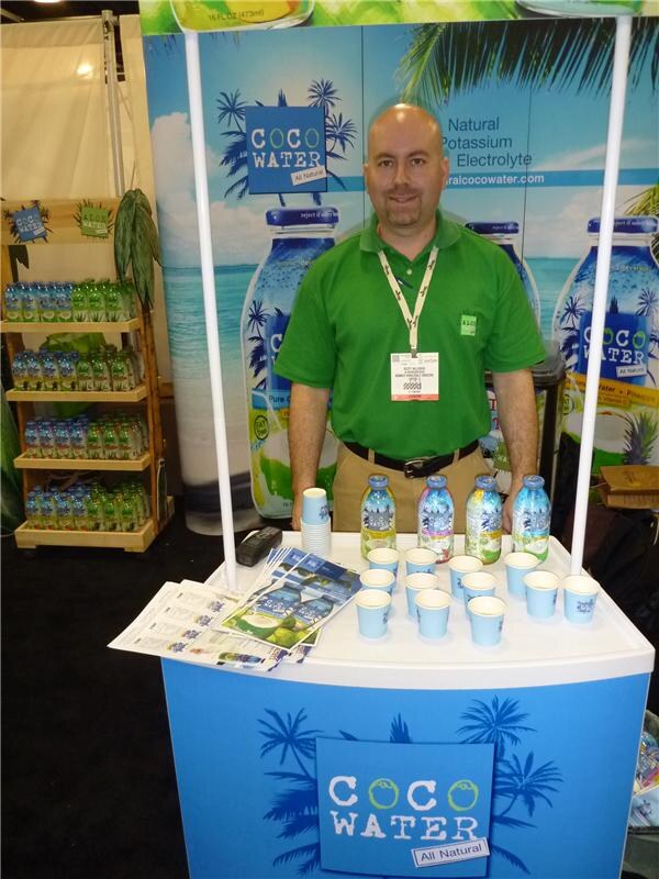 A bald man with a goatee in a green polo and show lanyard stands behind a Coco Water counter holding four bottles, small blue cups and brochures. Shelves of bottles are at left.