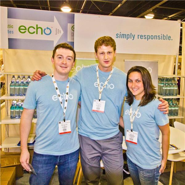 Two men and a woman in light blue Echo t-shirts and lanyards pose with arms around each other in front of shelves of bottled water and an Echo sign reading 'simply responsible.'