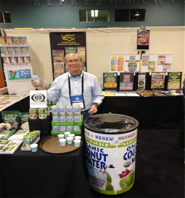 A man in a blue checked shirt holds a can at a booth with Edward & Sons packaged foods, a Non GMO Project sign and a large coconut water cooler in the foreground.