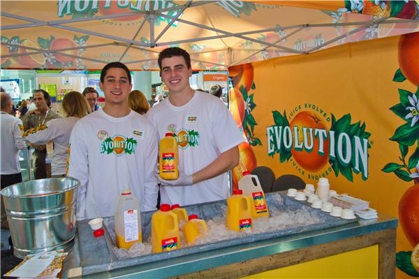 Two young men in white Evolution t-shirts stand behind a counter of iced orange juice bottles under an orange tent with Evolution orange graphics. Small cups sit beside them.
