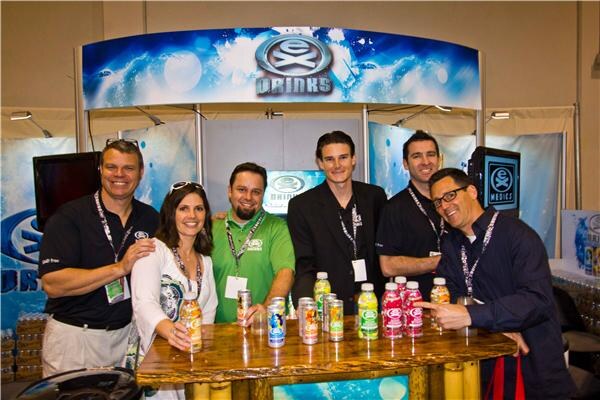 Six people, five men and one woman, stand behind a wooden bar table holding and displaying bottles and cans of EX Drinks beverages beneath an EX Drinks sign.