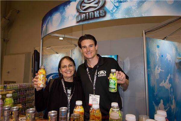 A smiling woman and a man in a black EX Drinks polo hold bottles of EX Drinks beverages in front of the curved EX Drinks booth sign. Cans and bottles sit on the counter below.