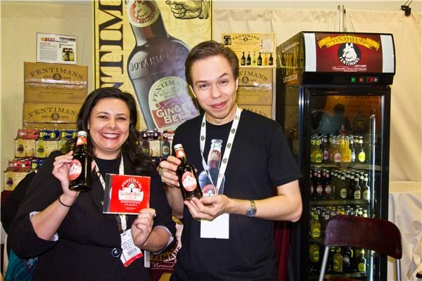 A smiling woman and a young man in a black t-shirt hold Fentimans bottles at a booth with stacked Fentimans cartons and a lit display fridge of bottled drinks behind them.