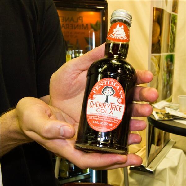 A close view of two hands holding a bottle of Fentimans Cherry Tree Cola with a red and white label, in front of a booth with a refrigerated case in the background.
