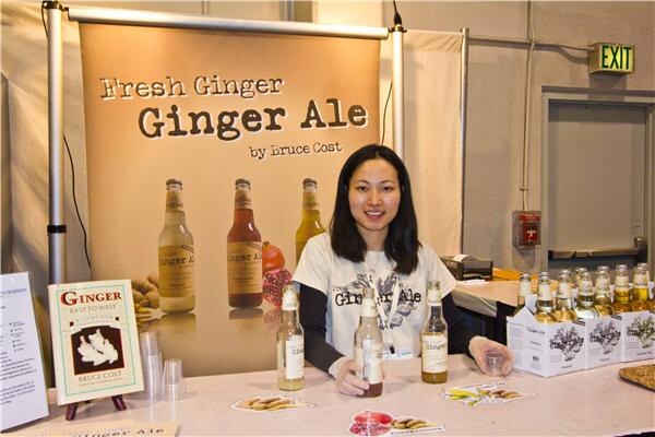 A woman in a graphic t-shirt stands behind a table with bottles of Fresh Ginger Ginger Ale by Bruce Cost, in front of a banner with the brand name and a Ginger book on a stand.