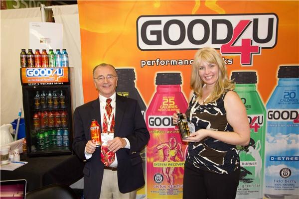 A man in a dark blazer and a blonde woman hold Good4U drink bottles in front of a large orange Good4U banner showing performance drink bottles and a small Good4U display cooler.