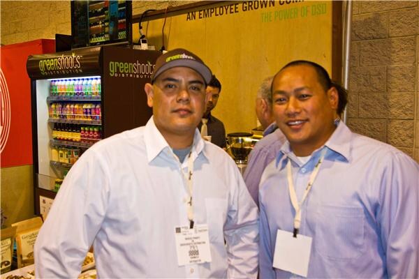 Two men wearing event badges pose in front of a GreenShoots Distribution refrigerated display of bottled drinks; one wears a cap, and other attendees stand behind them.