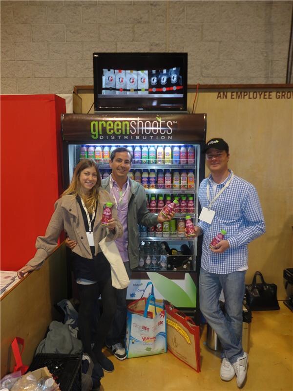 A woman and two men hold bottles of pink drink beside a Green Shoots Distribution refrigerated display case filled with bottled beverages, with shopping bags at their feet.