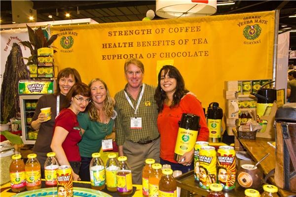 Four people pose at the Guayaki booth in front of a yellow banner reading strength of coffee, health benefits of tea, euphoria of chocolate, with Guayaki bottles and cans in the foreground.