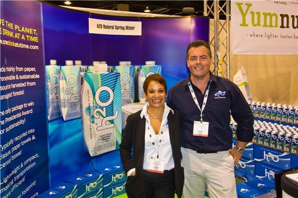 A woman in a black blazer and a man in a navy shirt stand together at the h2o Natural Spring Water booth, with h2o cartons displayed behind them and a Yumnuts banner overhead.