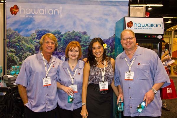 Four people in a row, three in light blue Hawaiian Springs shirts and a woman with a flower in her hair, hold water bottles in front of a Hawaiian Springs banner showing a green landscape.
