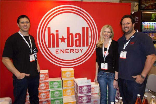 Two men and a blonde woman stand in front of a red Hi Ball Energy sign beside stacked cases of the drink in several colors, all wearing event badges.