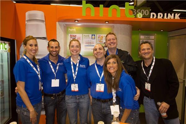 Six people, most in blue Hint polo shirts with lanyards, crowd together at the orange and green Hint booth, smiling; one man in a black jacket holds a bottle.