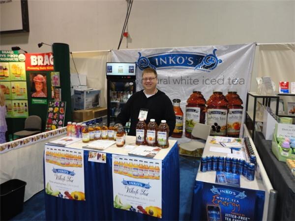 A man in a black sweater stands behind a booth table of Inko's white iced tea bottles under an Inko's banner. A Bragg display is to the left and Inko's White Tea Energy cans are at right.
