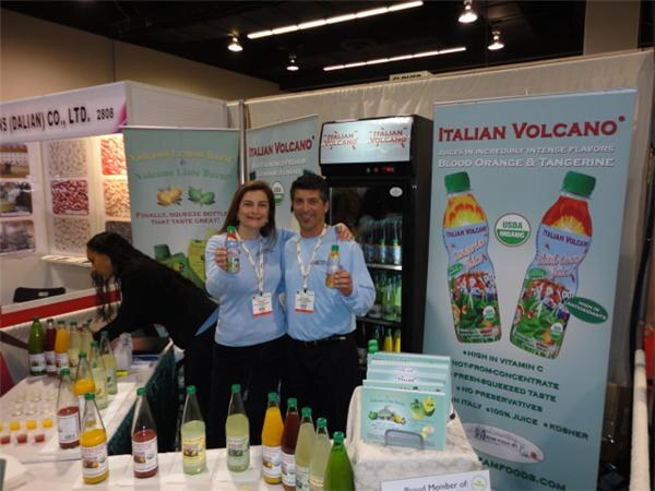 A woman and a man in light blue shirts and lanyards hold bottles in front of an Italian Volcano banner for blood orange and tangerine juice. Juice bottles line a table in front of them.