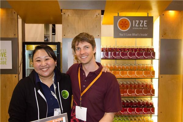 A smiling woman in a dark hooded jacket and a smiling man in a maroon shirt with an orange lanyard stand in front of a lit shelf of Izze sparkling juice bottles and an Izze sign.