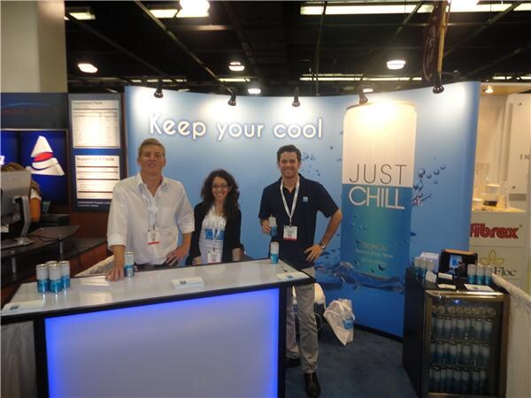 Two men and a woman stand at a lit booth counter with Just Chill cans on it. A blue backdrop reads Keep your cool, and a Fibrex sign is visible at right.