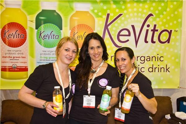 Three women in black shirts with show badges smile and hold bottles of KeVita probiotic drink in front of a green and yellow KeVita banner showing bottles.