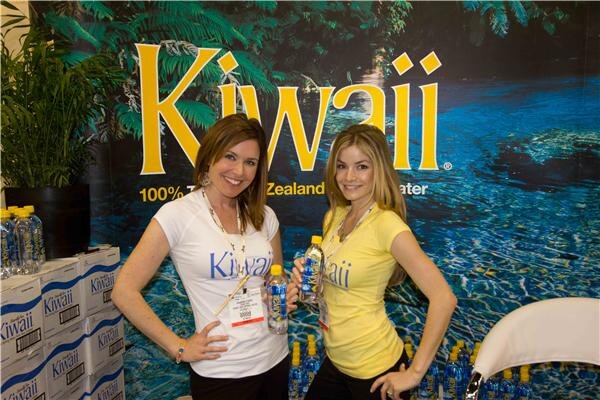 Two smiling women in Kiwaii shirts hold bottles of water in front of a large Kiwaii banner showing a blue pool, with cases of Kiwaii water stacked at left.