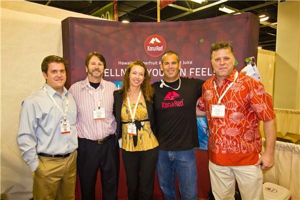 Five people stand side by side in front of a Kona Red banner. One man wears a black Kona Red shirt, another an orange patterned shirt, and the rest wear show badges.