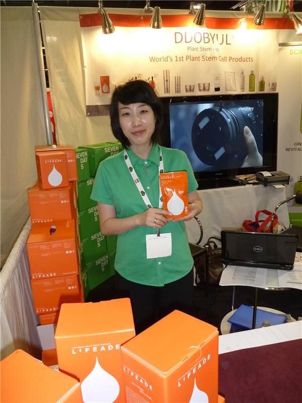 A woman in a green shirt holds an orange Lifeade pouch beside stacked orange Lifeade boxes and green Seven Days boxes, with a DDOBYUL sign and a TV behind her.