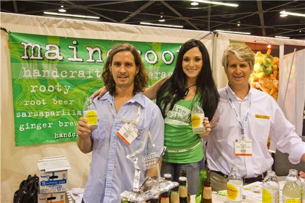 Two men and a woman with long dark hair pose arm in arm in front of a green Maine Root banner. Two hold bottles of soda, and a trophy and more bottles sit on the table in front.