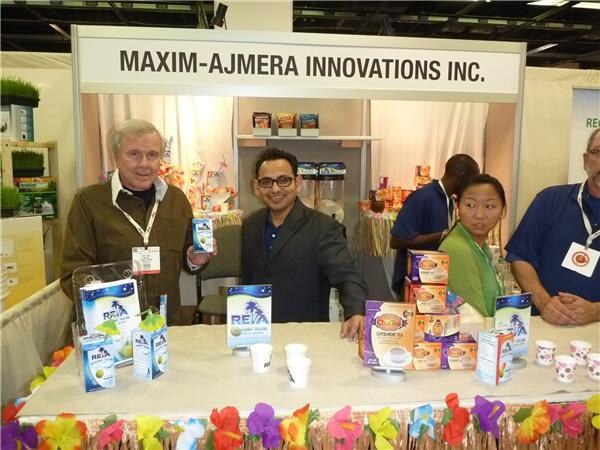 Two men stand behind a flower-lei-trimmed table at the Maxim-Ajmera Innovations Inc. booth, with Rea boxes and cups on display. Two more people stand in the background.