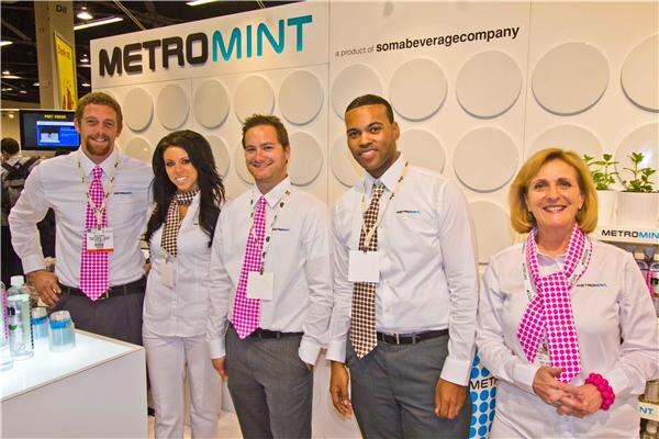 Five smiling people in white shirts and pink or brown patterned ties and scarves stand in front of a white Metromint wall, which reads a product of soma beverage company.