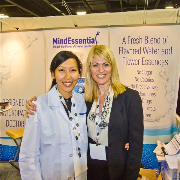 Two smiling women stand with arms around each other in front of MindEssential banners. One wears a white lab coat, the other a dark blazer. A banner reads A Fresh Blend of Flavored Water and Flower Essences.