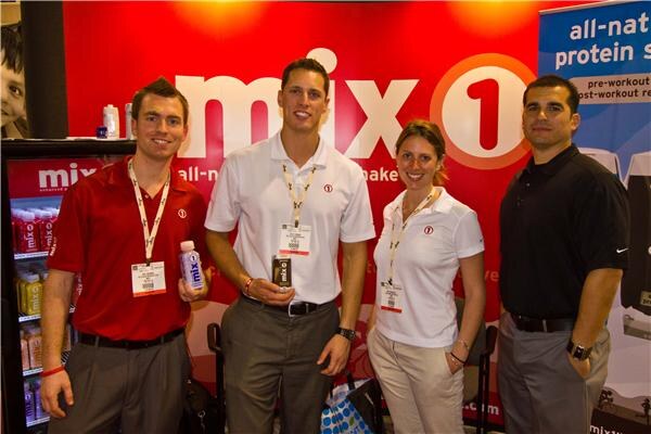 Three men and a woman stand in front of a red Mix1 backdrop. Two of the men hold Mix1 drink containers, and a refrigerated display of drinks sits at left.