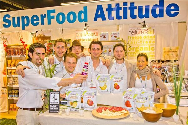 Seven people crowd together behind a table of freeze-dried fruit packages under a SuperFood Attitude banner, with GojiAnia and TerrAmazon signs behind them.