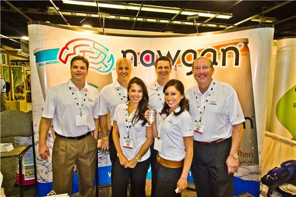 Six people in white polo shirts, four men and two women, stand in front of a curved Nawgan display with a brain logo. One woman holds a can.