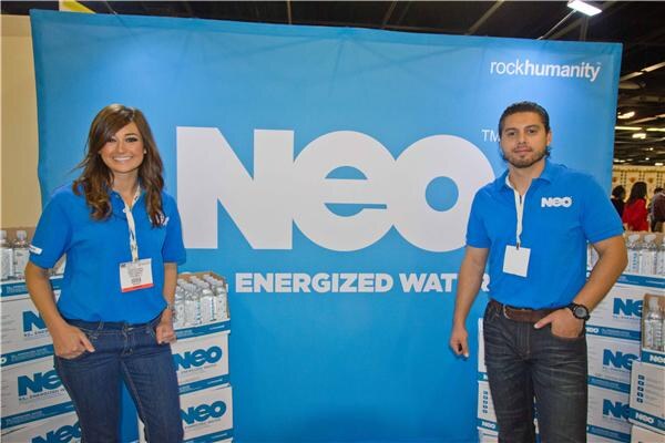 A woman and a man in blue NEO polo shirts stand in front of a blue NEO Energized Water backdrop with rockhumanity text, with cases of bottled water stacked behind them.