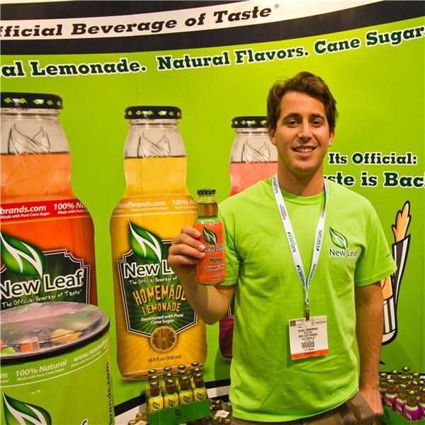 A smiling man in a green New Leaf T-shirt and a show badge holds up a bottle of New Leaf tea in front of a green backdrop with large New Leaf bottles, including Homemade Lemonade.