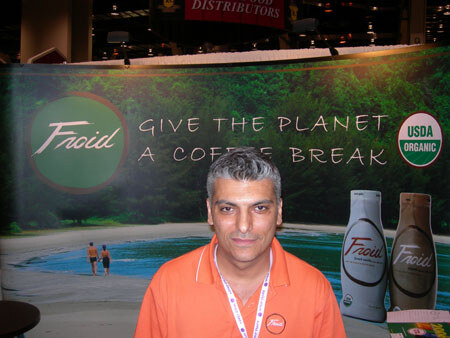 A man in an orange polo and lanyard stands in front of a Froid backdrop reading "Give the planet a coffee break," with USDA Organic seal and two Froid bottles.