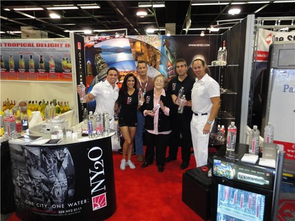 Six people stand together on red carpet in the NY2O booth, beside a black NY2O counter reading One City, One Water, with bottled water on shelves and a display backdrop behind them.