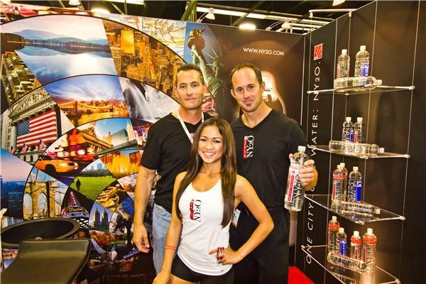 Two men in black NY2O shirts and a woman in a white NY2O tank top pose in front of a city-themed NY2O backdrop, one man holding a bottle, with bottles on glass shelves beside them.