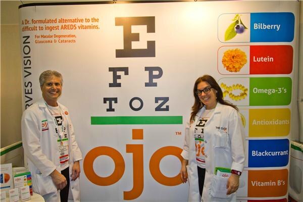 A man and a woman in white lab coats stand in front of an ojo display with an eye chart and a list of ingredients including Bilberry, Lutein and Omega-3's.