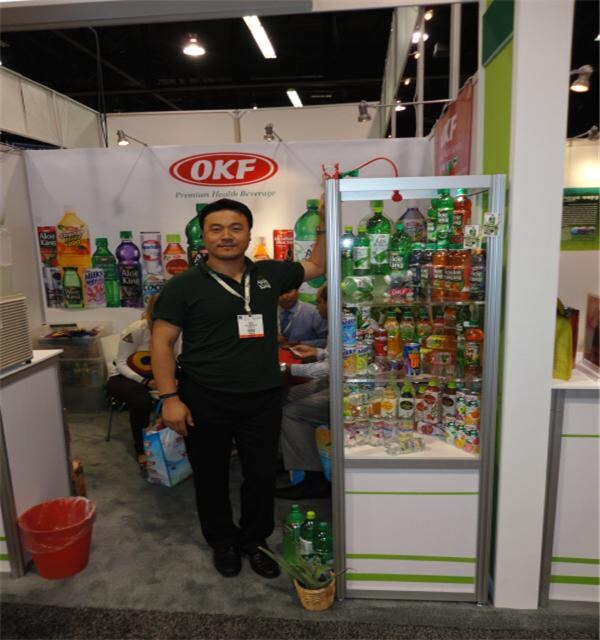 A man in a dark green polo stands beside a glass display case of OKF beverage bottles, in front of an OKF Premium Health Beverage banner.