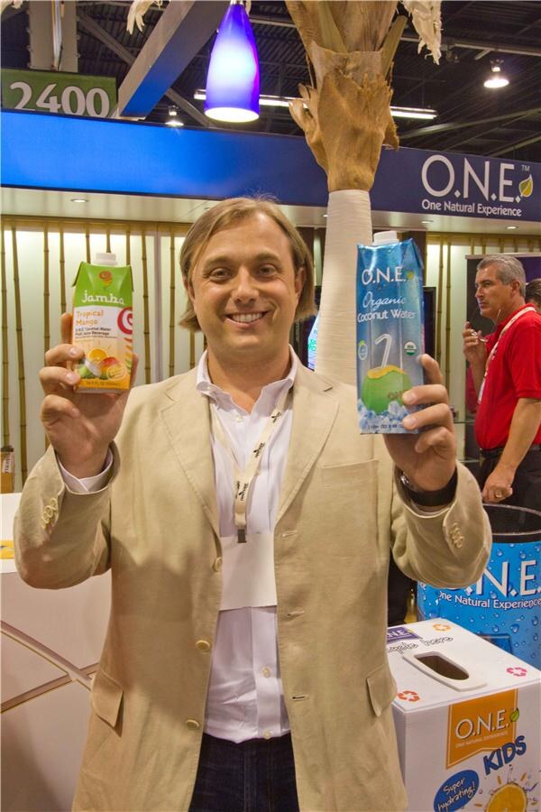 A smiling man in a beige blazer holds up a Jamba Tropical Mango coconut water carton and an O.N.E. Organic Coconut Water carton in front of the O.N.E. One Natural Experience booth.