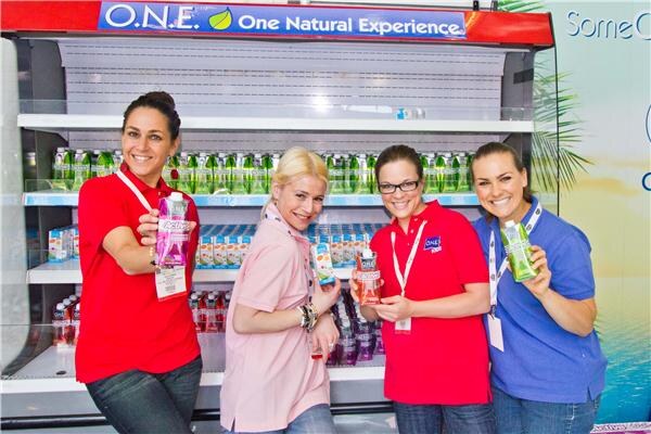 Four smiling women in red, pink and blue tops hold O.N.E. coconut water cartons in front of a refrigerated display case under an O.N.E. One Natural Experience sign.
