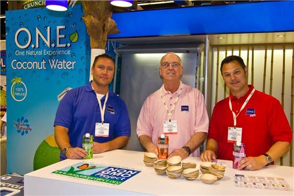 Three men in blue, pink and red shirts stand behind a counter holding O.N.E. cartons, with small bowls on the counter and an O.N.E. Coconut Water sign behind them.