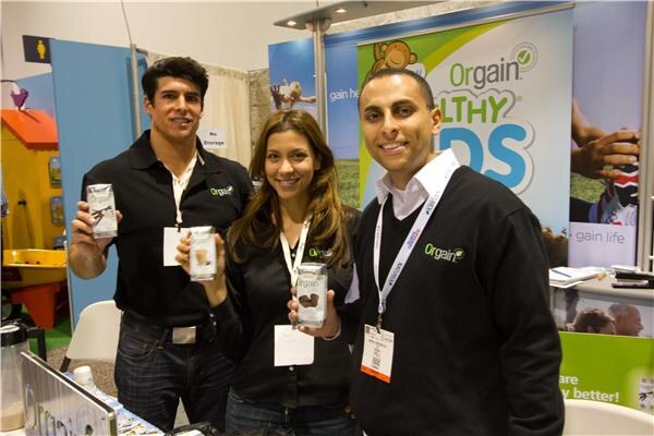 Two men and a woman in dark Orgain shirts smile at the camera, the man and woman at left holding Orgain cartons, in front of an Orgain Healthy Kids banner.