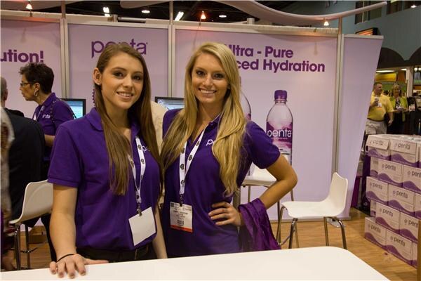 Two smiling women in purple polo shirts and lanyards stand at a Penta booth with purple banners, a Penta water bottle graphic and stacked Penta cases behind them.