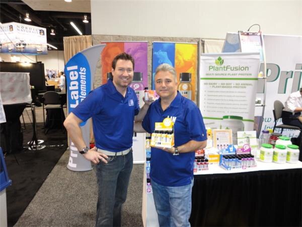 Two men in blue polo shirts stand at a booth table; one holds a small box of bottled shots. A PlantFusion plant protein banner and tubs of powder are behind them.