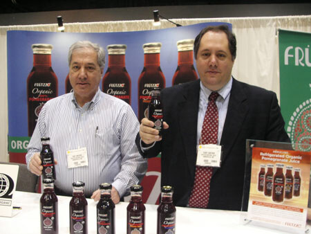 Two men with name badges stand behind a table of Frutzzo organic pomegranate juice bottles; one holds a bottle, with a Frutzzo banner and product sign behind them.