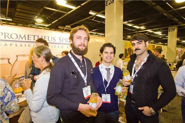 Three men with lanyards hold bottles of yellow drink in front of a Prometheus Springs banner, with a woman and other attendees behind them and glass jars on the left.
